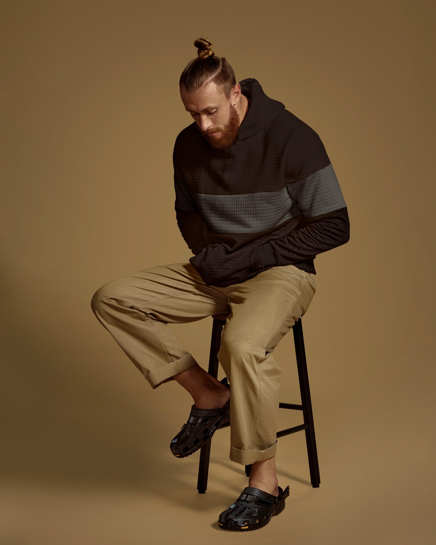 Adult male with a beard and a top knot sits on a stool wearing a hoodie, beige pants, and clogs in a studio setting with a neutral background.